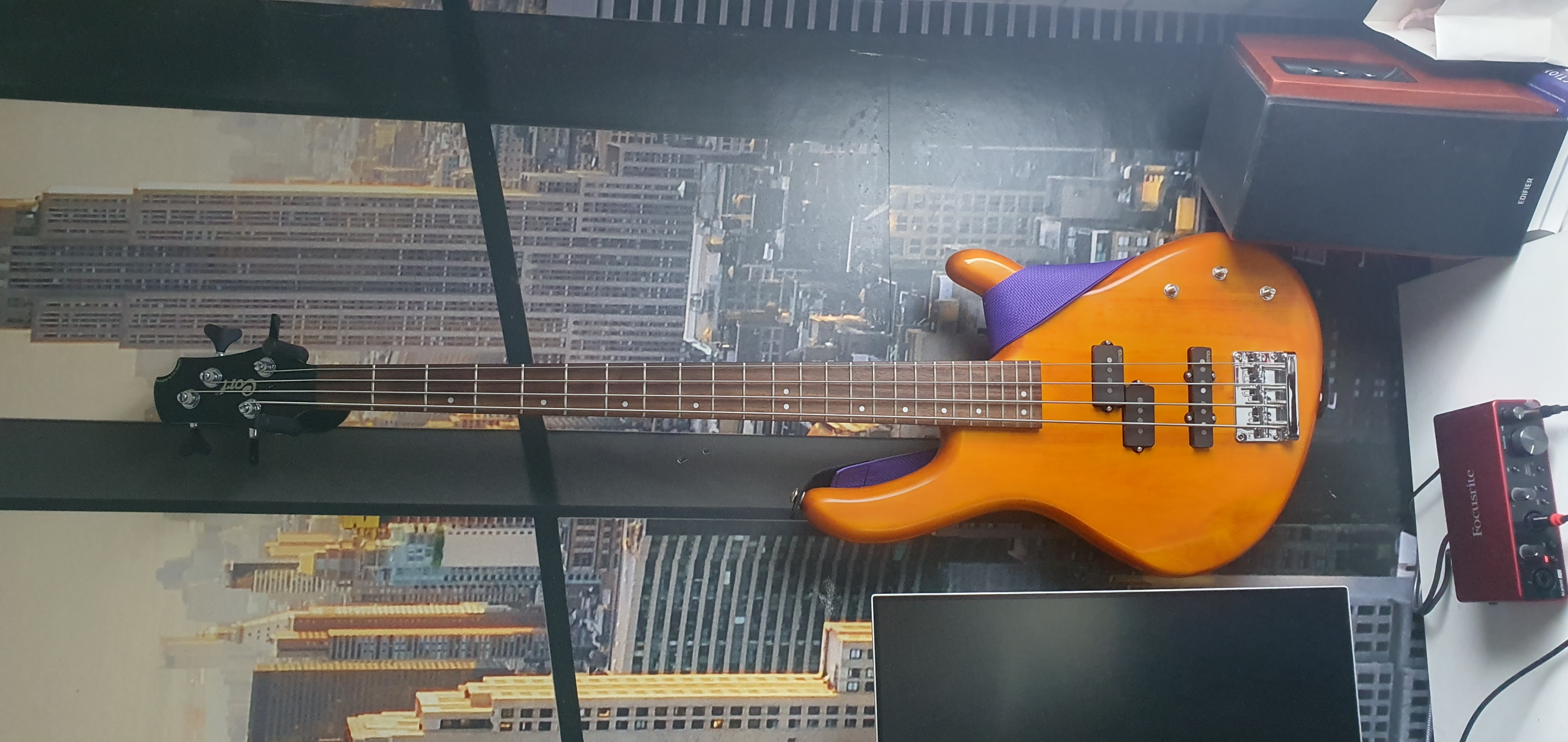 A Cort Action PJ hanging on the wall, it has a purple strap and the bass has a glossy almost orange wood